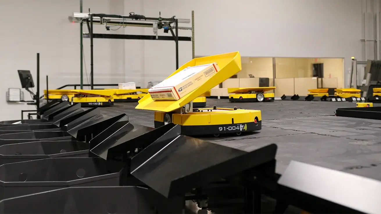 tSort autonomous mobile robots carrying tilt trays across a sorting platform, one tilting to discharge a parcel into a chute below.